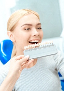 A young female with blonde hair looking at her teeth in the mirror while a dentist holds up a shade guide