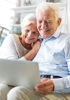 An older couple seated on a couch using a laptop to converse with family and smiling after receiving full-mouth reconstruction in Marble Falls