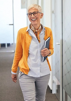 An older woman wearing business casual clothing and smiling as she walks down the hallway of her office